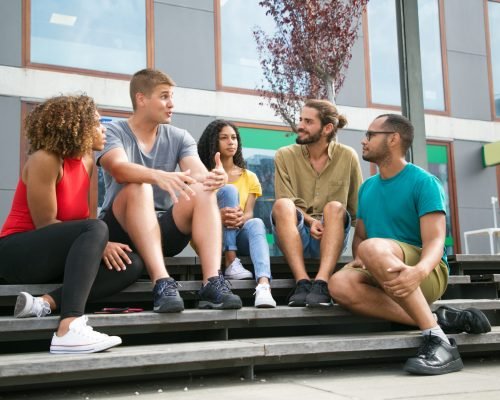 Joyful Caucasian guy telling exciting story to friends. Interracial group of young people sitting on outdoor staircase, talking, listening, laughing. Happy friends concept